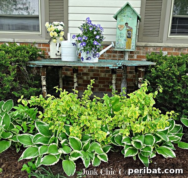 A weathered wooden table styled with old white watering cans and rustic birdhouses, all filled with flowers, in a garden corner