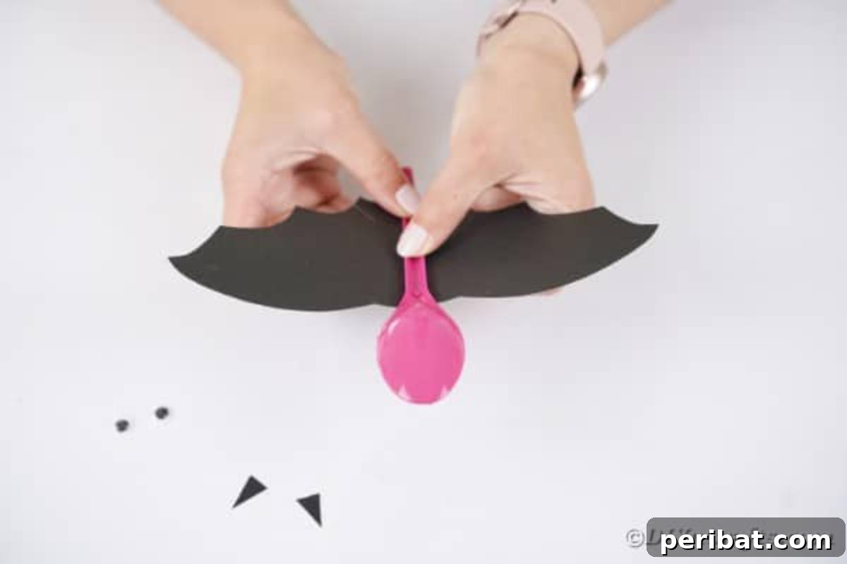 Adorable Recycled Plastic Spoon Bat Halloween Decor 13 A spoon being glued onto the center of black paper bat wings, illustrating the primary step of assembling the upcycled Halloween craft.
