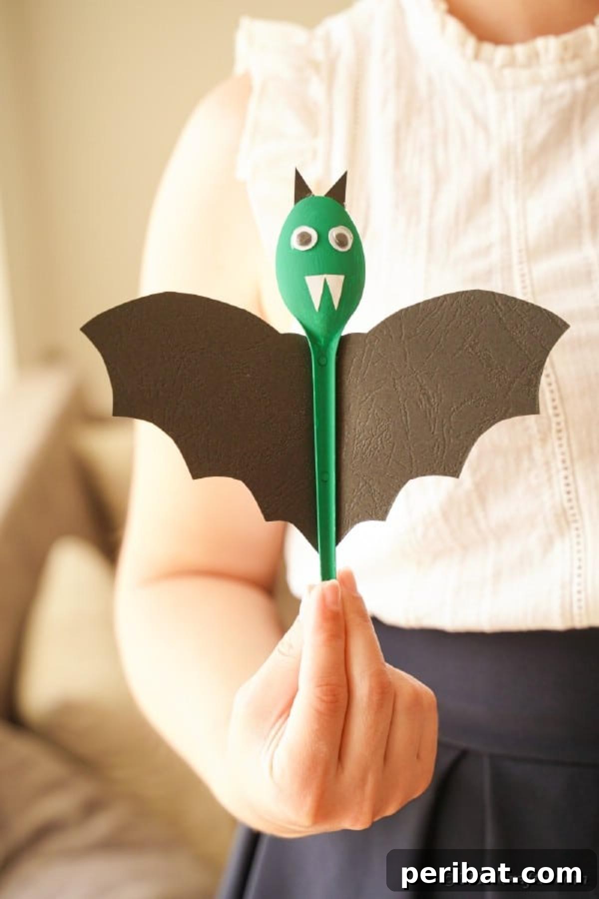 Adorable Recycled Plastic Spoon Bat Halloween Decor 18 A person holding an upcycled spoon bat upright, as if flying, against a plain background, highlighting its painted body and paper wings for Halloween.