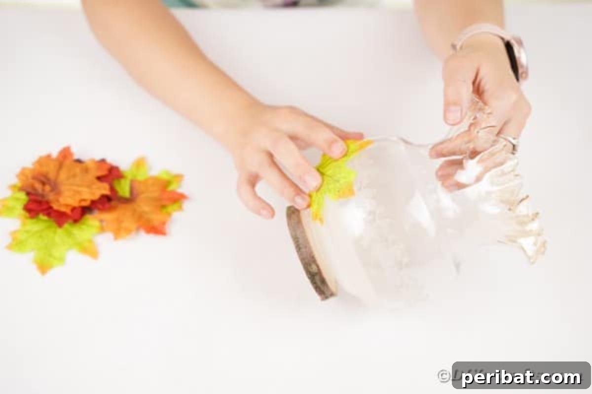 Autumn Leaf Jar Luminaries 10 A hand carefully applies glue to an artificial fall leaf before pressing it onto the exterior of a glass jar, demonstrating the leaf placement step.