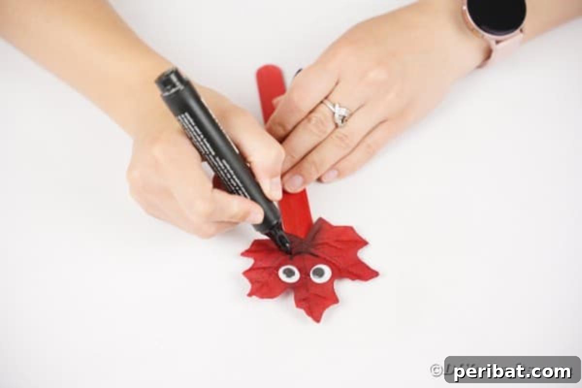 Geri Dönüşümle Sonbahar Yaprak İnsanlarını Canlandırın 13 A hand using a black marker to draw a smile onto the artificial leaf head of a leaf person craft, completing its facial features.