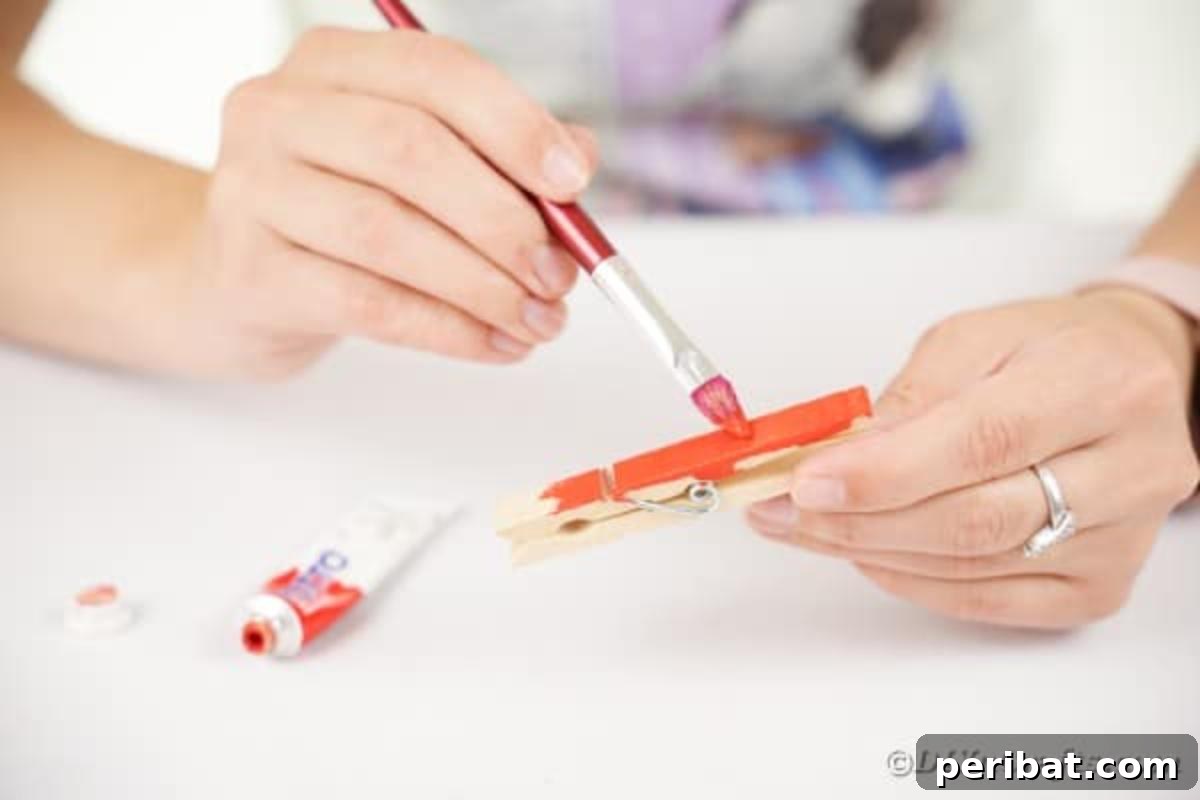 Geri Dönüşümle Sonbahar Yaprak İnsanlarını Canlandırın 8 A hand holding a clothespin covered in orange paint, illustrating the first step of painting the craft bases for leaf people.