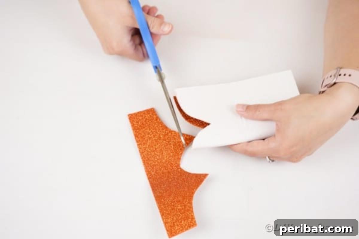 A hand carefully cutting an owl face shape from orange glitter paper, which will form the top part of the owl's head.
