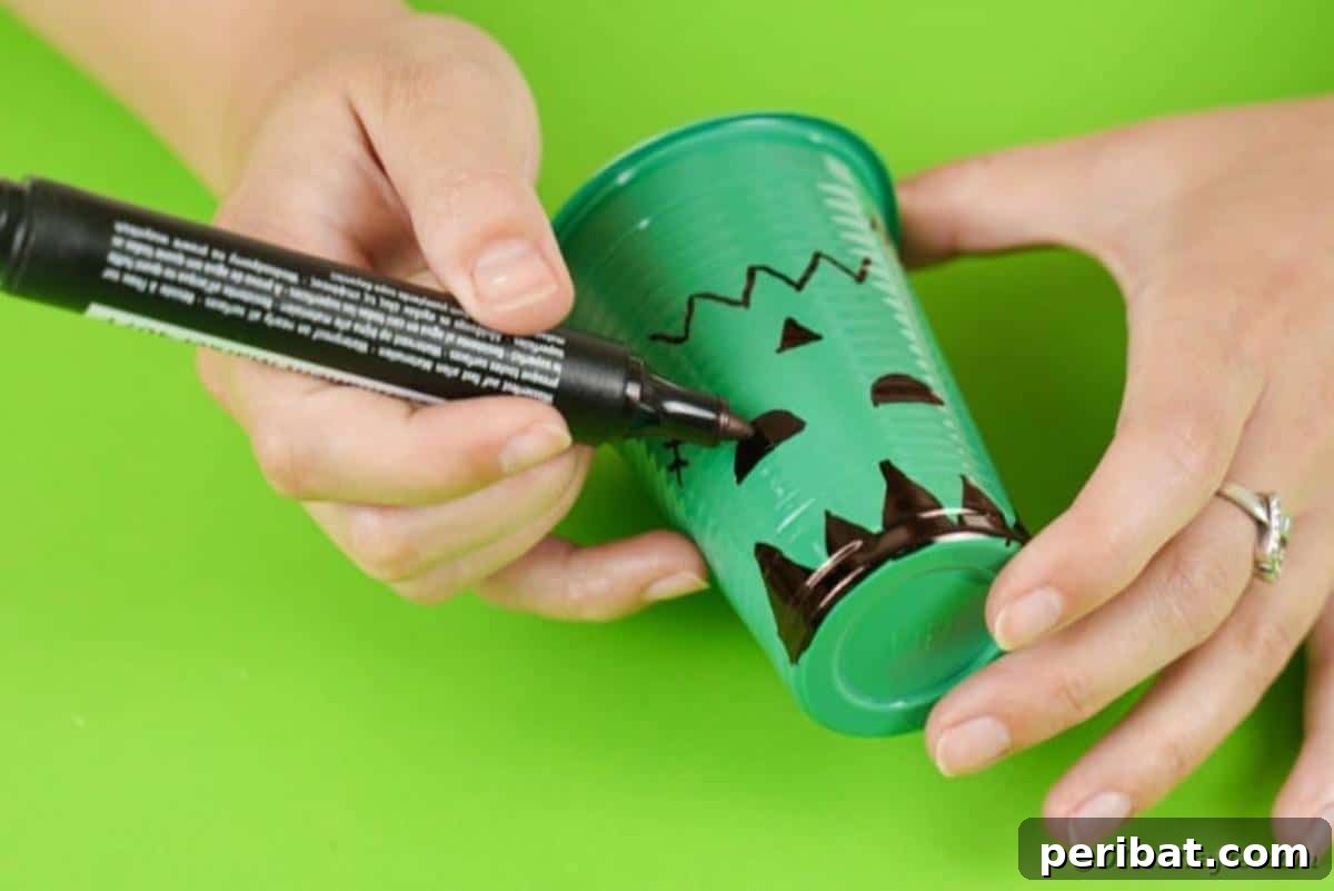 Close-up of a hand drawing the distinctive face of Frankenstein onto a green plastic cup.