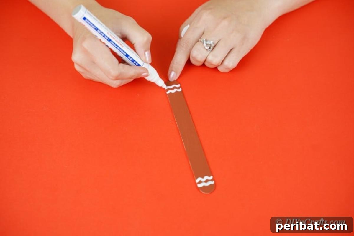 Sweet and Snowy Craft Stick Holiday Decor 10 Applying white paint details onto a brown popsicle stick for a gingerbread man ornament