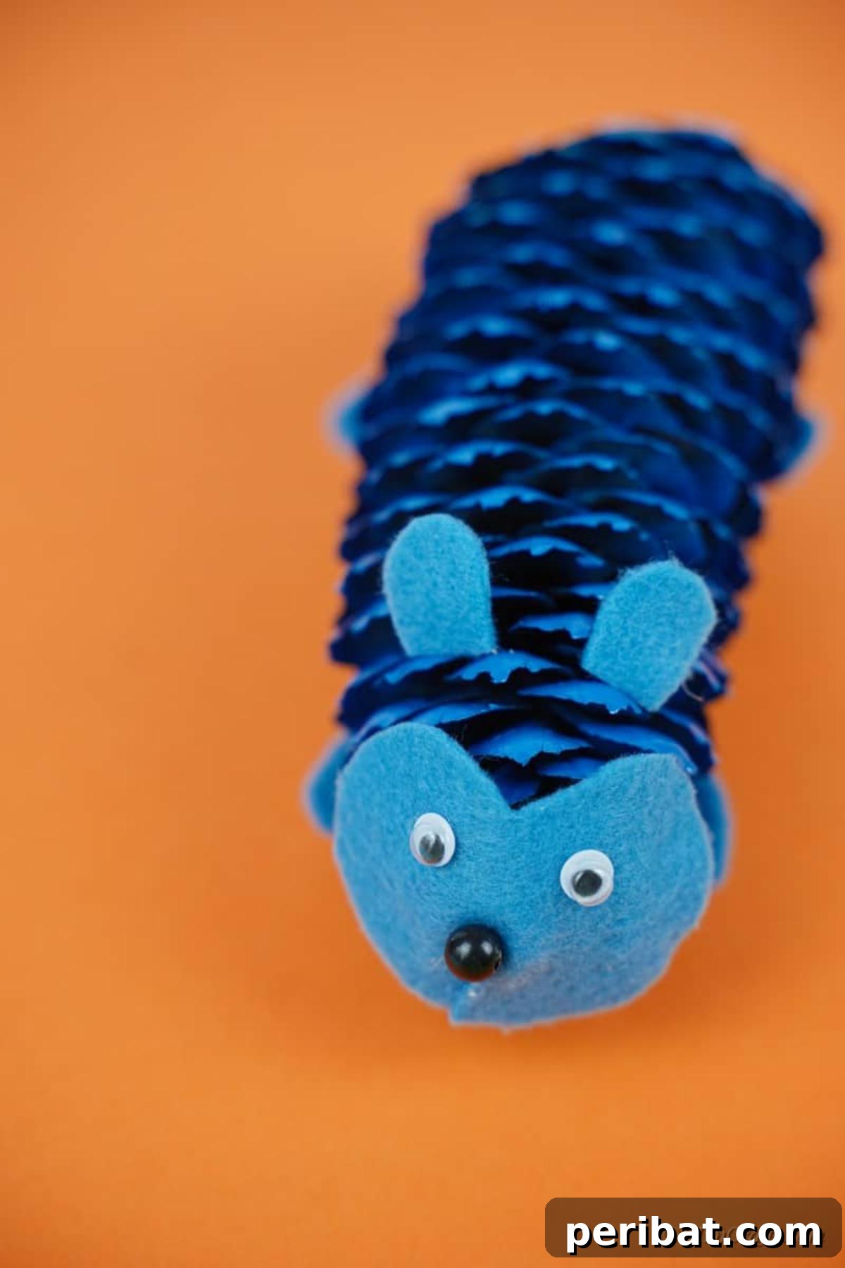 Whimsical Pinecone Hedgehogs 17 A single blue pinecone hedgehog craft sitting on a vibrant orange paper surface, highlighting its painted details and cute design.