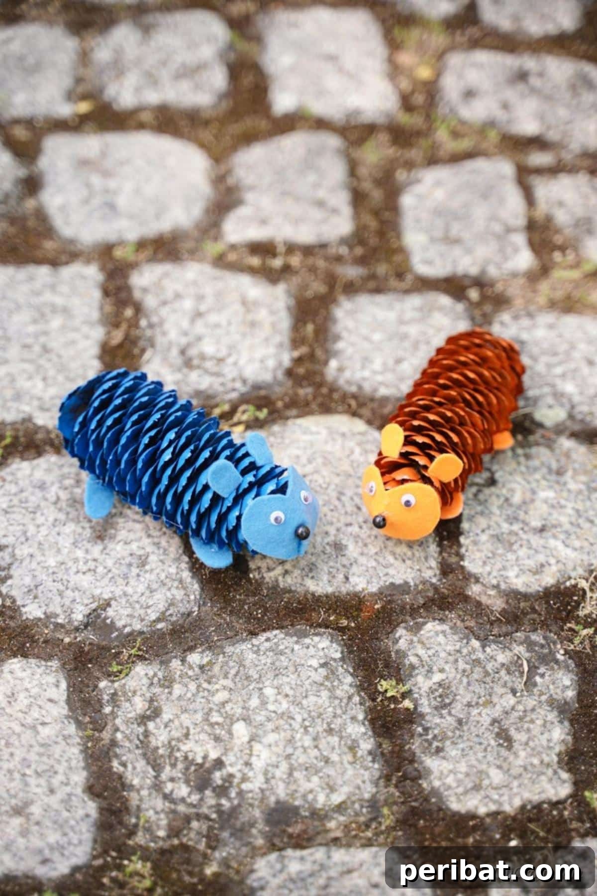 Whimsical Pinecone Hedgehogs 5 Two painted pinecone hedgehogs, one blue and one orange, sitting on cobblestone pavers amidst fallen autumn leaves, ready for display.