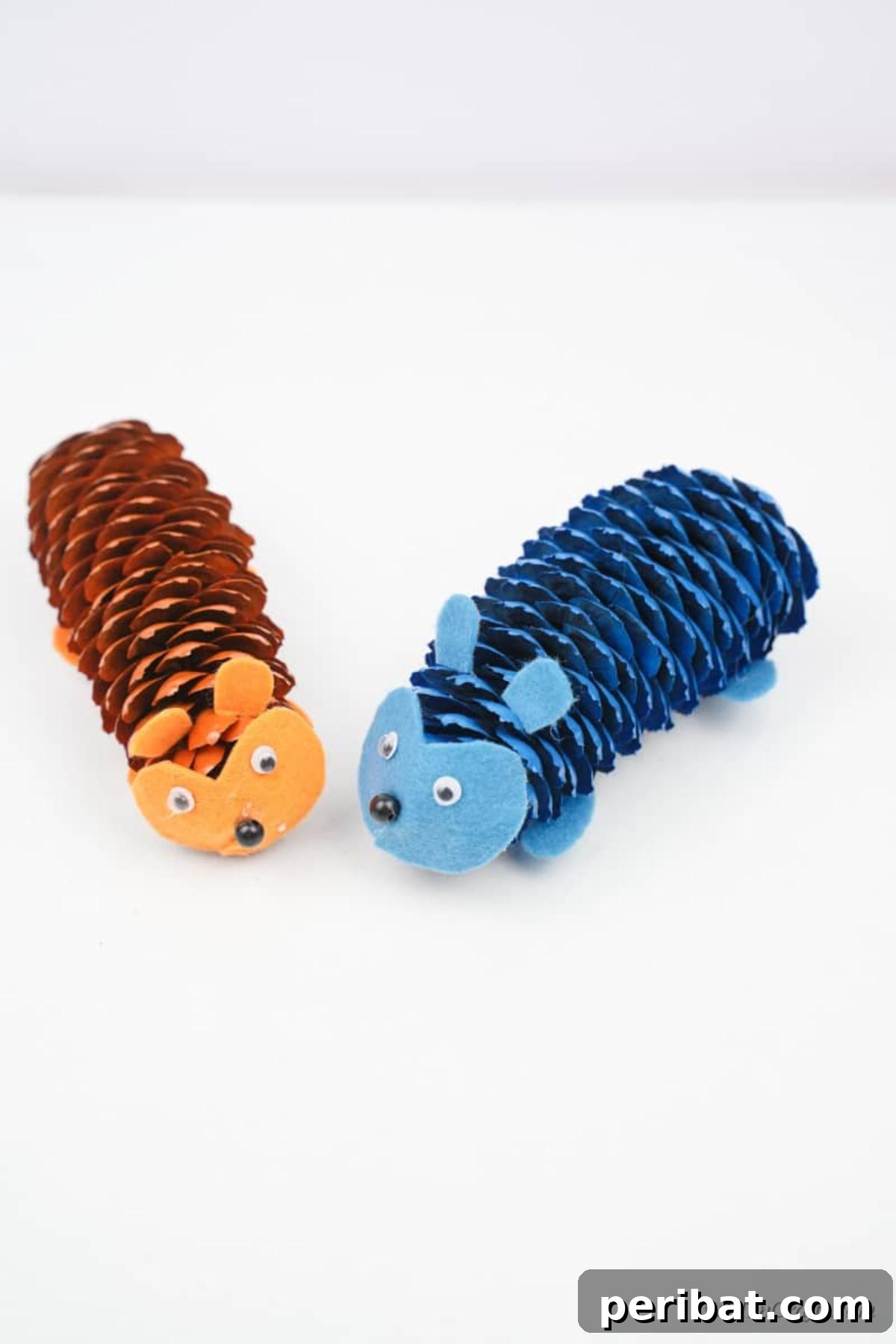 Whimsical Pinecone Hedgehogs 7 Two charming pinecone hedgehogs, one blue and one orange, arranged on a clean white table with scattered autumn leaves, showcasing their vibrant colors and simple design.