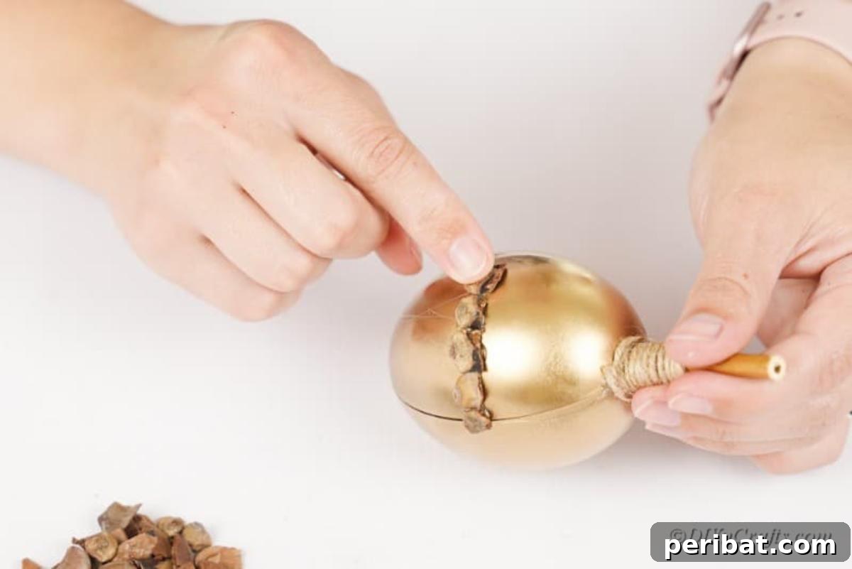 Atık Plastik Yumurtalardan Sonbahar Meşe Palamudu Yapımı 12 Carefully placing small pinecone pieces onto a golden craft acorn to form its textured cap, on a white table.