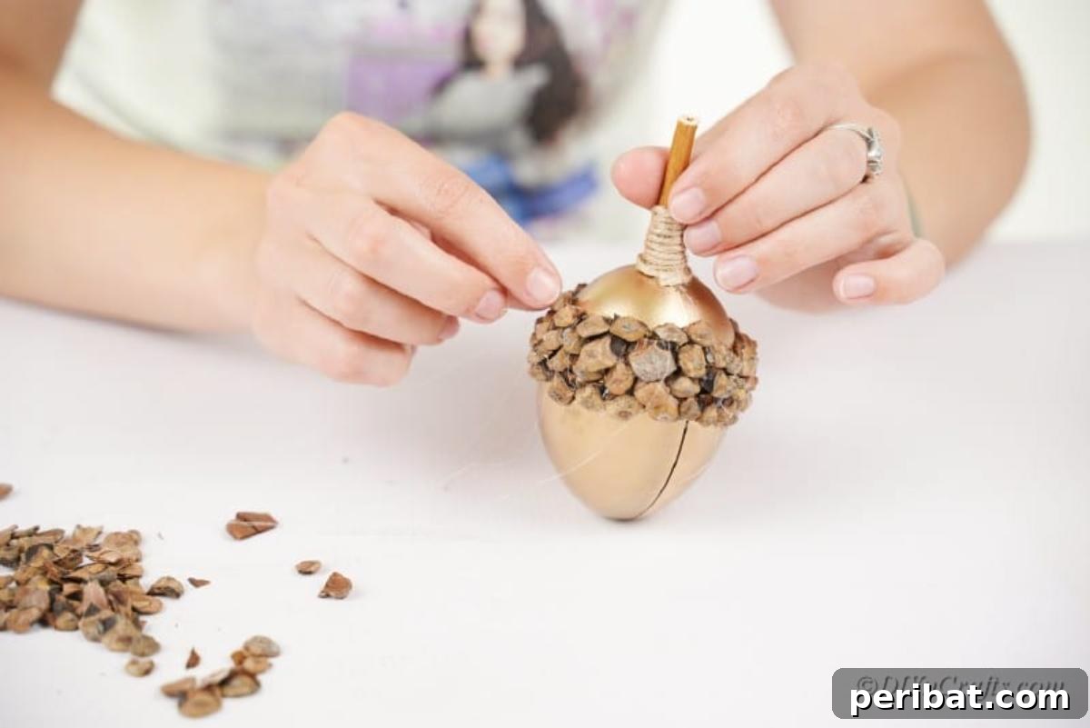 Atık Plastik Yumurtalardan Sonbahar Meşe Palamudu Yapımı 13 Detailed shot of small pinecone pieces being arranged and glued onto a golden acorn craft to create an intricate cap design.