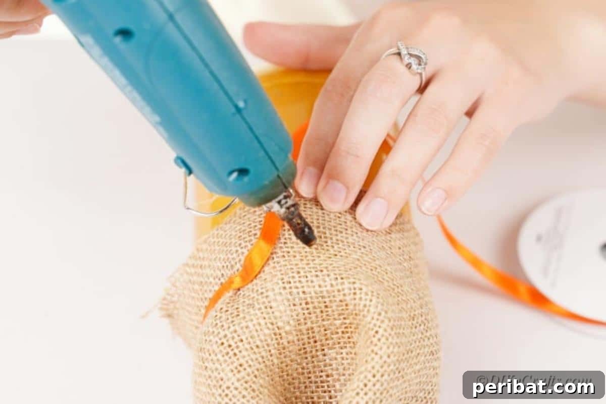 Whimsical Fall Scarecrow Jar Craft 11 A hot glue gun secures an orange ribbon around the burlap hat of a scarecrow jar, adding a decorative band to the craft.