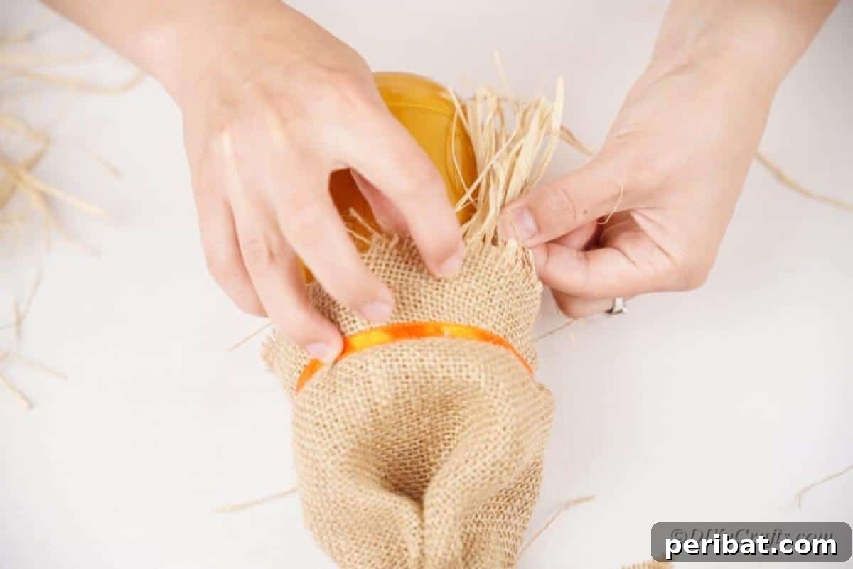 Whimsical Fall Scarecrow Jar Craft 12 Crafting the scarecrow's hair: Straw pieces are being inserted into the hot glue under the burlap hat of the jar, creating a rustic and charming effect.