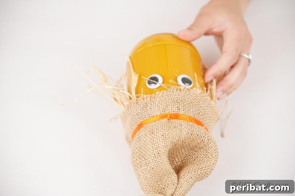 Whimsical Fall Scarecrow Jar Craft 13 Googly eyes are glued onto the painted jar, sitting beneath the burlap hat and straw hair, bringing the scarecrow craft to life.