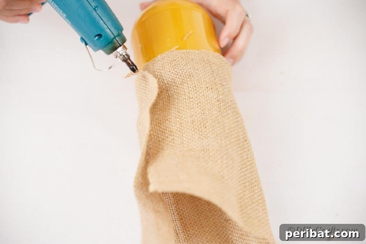Whimsical Fall Scarecrow Jar Craft 9 A hot glue gun is used to carefully apply glue to a piece of burlap, securing it around the top of a jar to form the base of the scarecrow's hat.