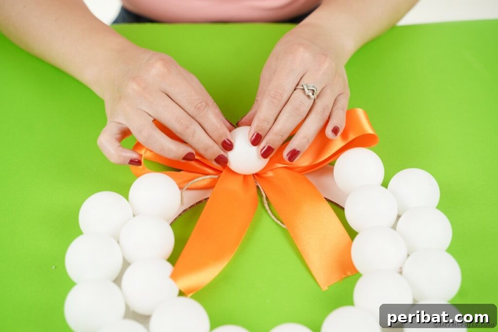 Ghoul's Eye Halloween Entry Wreath 15 A final ping pong ball being glued into the center of the orange ribbon bow, creating a central 'eyeball' focal point on the wreath