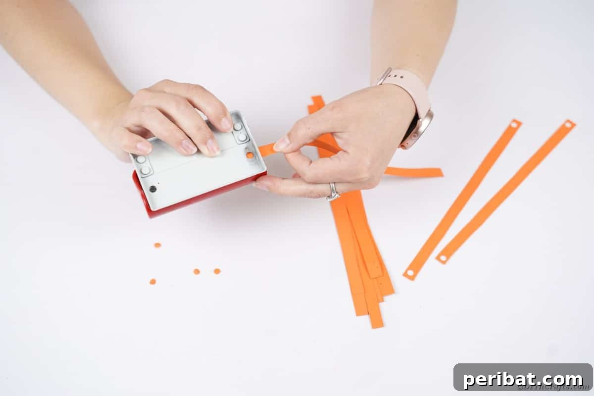 Spooky Paper Strip Creations: Bats, Pumpkins & Spiders 11 A hand using a hole punch to create holes at the ends of several orange paper strips.