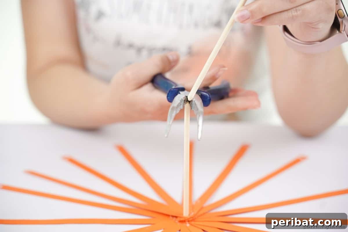 Spooky Paper Strip Creations: Bats, Pumpkins & Spiders 13 A hand holding a dowel rod, with orange paper strips attached, is about to be cut short.