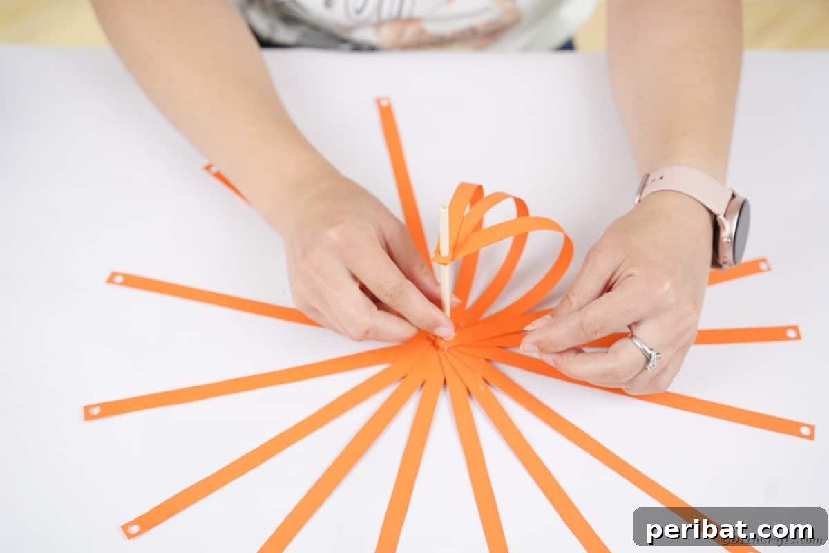 Spooky Paper Strip Creations: Bats, Pumpkins & Spiders 14 A hand gluing the top end of an orange paper strip to the dowel, creating the pumpkin's curve.