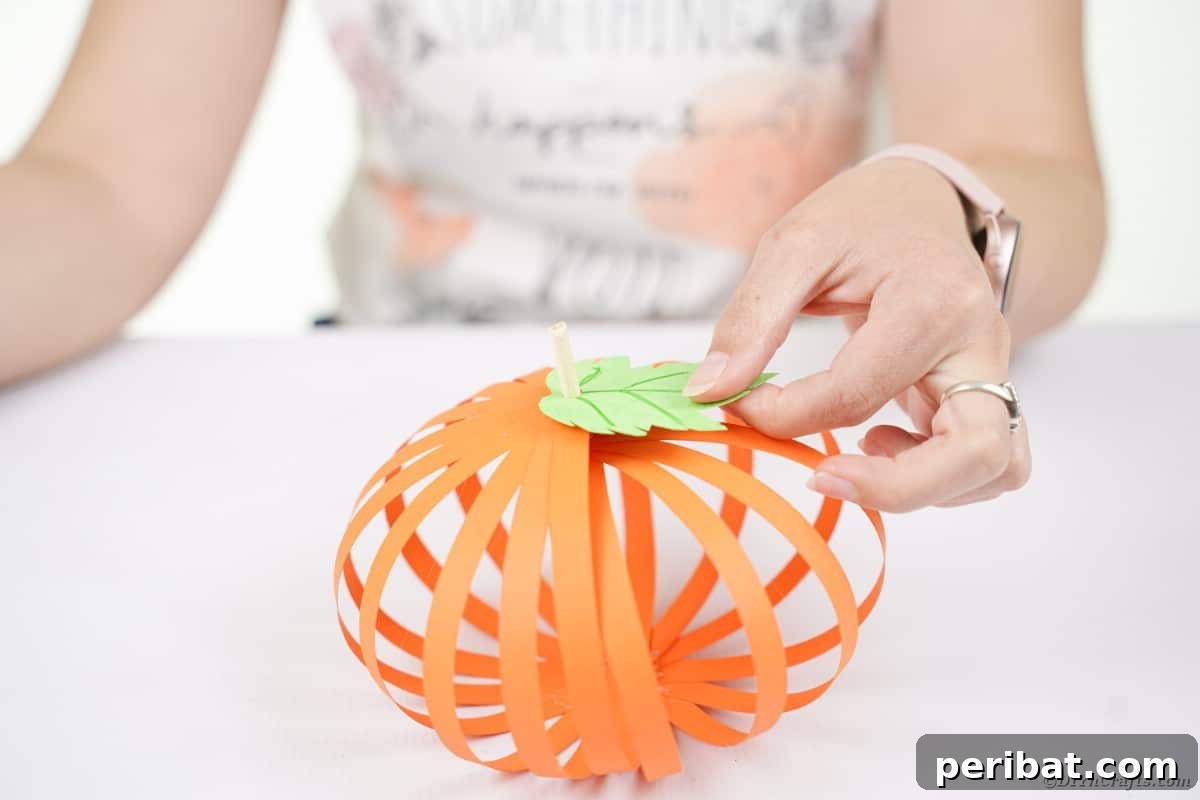 Spooky Paper Strip Creations: Bats, Pumpkins & Spiders 18 A green paper leaf is being glued onto the top of the orange paper pumpkin, around the dowel stem.