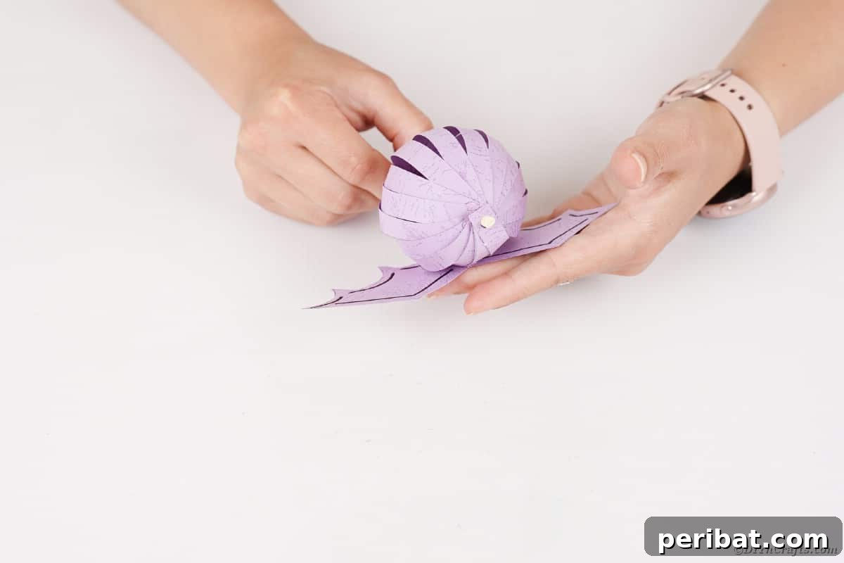 Spooky Paper Strip Creations: Bats, Pumpkins & Spiders 25 A pair of purple paper bat wings are being glued to the center of the rounded paper bat body.
