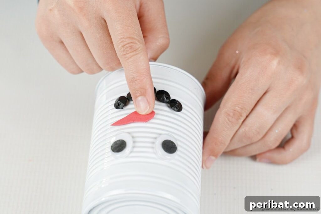 DIY Painted Tin Can Snowman Video Tutorial 22 Carefully gluing the orange foam carrot nose onto the snowman's face, positioning it between the eyes and mouth.