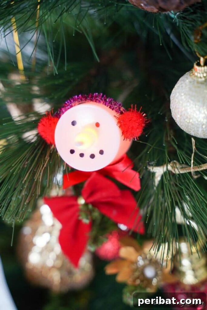 A festive snowman ornament made from an LED tealight candle, featuring painted facial details and a tiny knit hat, hanging from a branch.