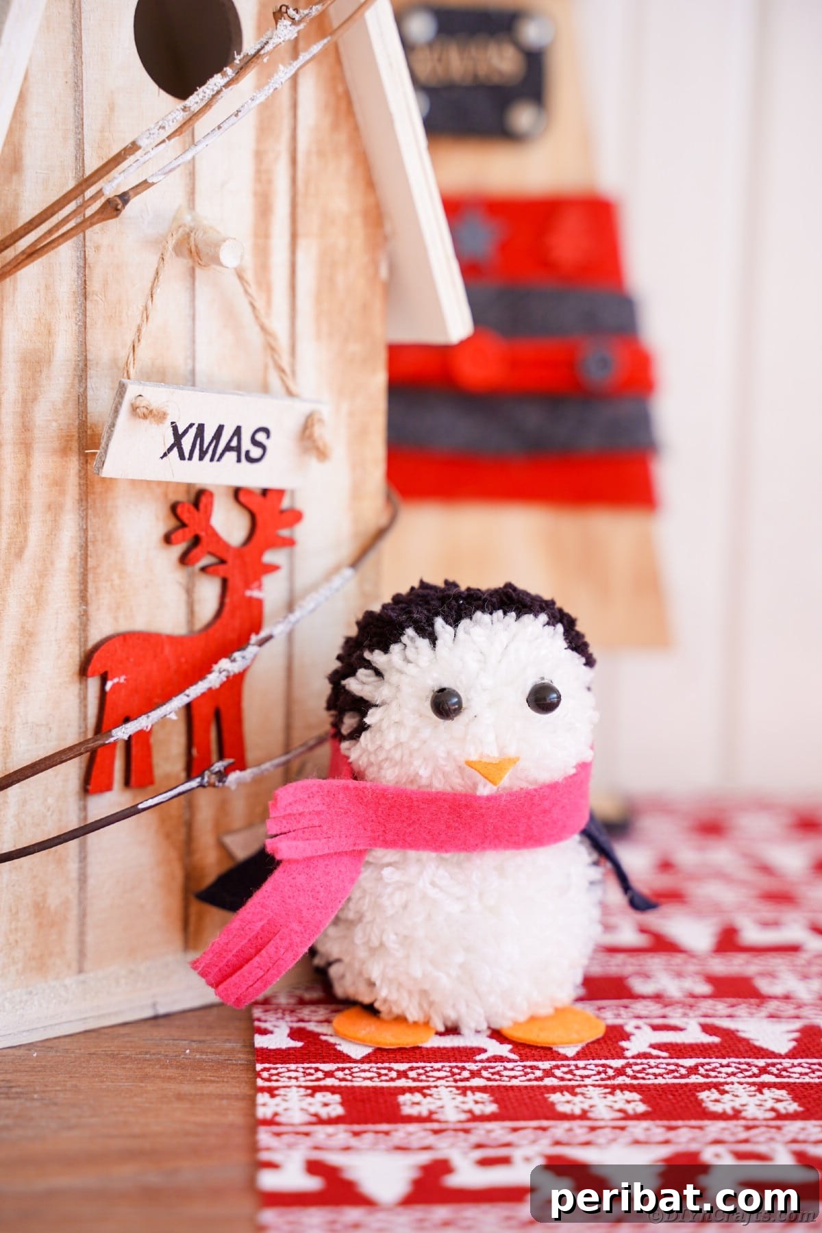 Handmade Pom Pom Penguin Winter Charm 23 Two pom pom penguins sitting on wooden winter decor, one with a red scarf, one with a pink scarf