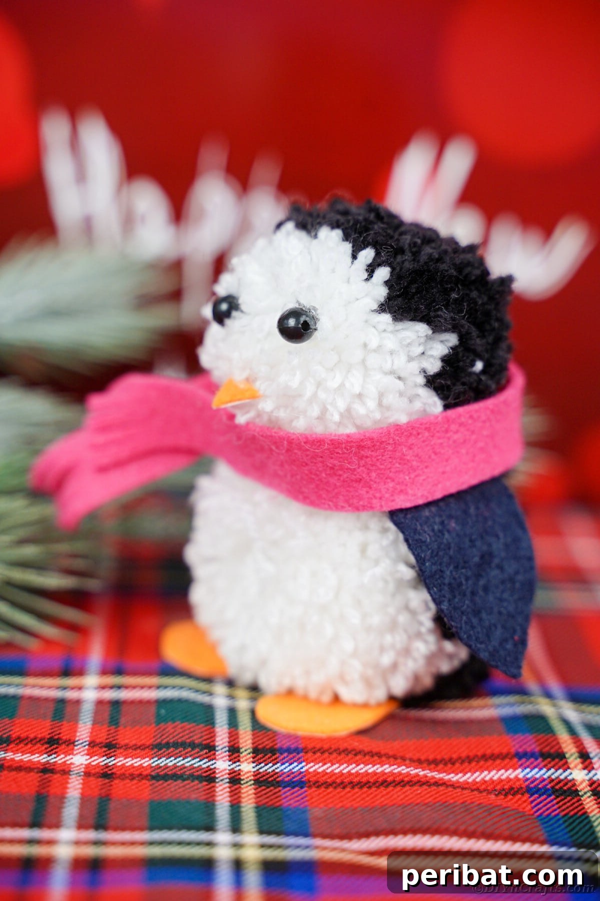 Handmade Pom Pom Penguin Winter Charm 25 Close-up of a pom pom penguin with a red scarf resting on a red and white flannel background