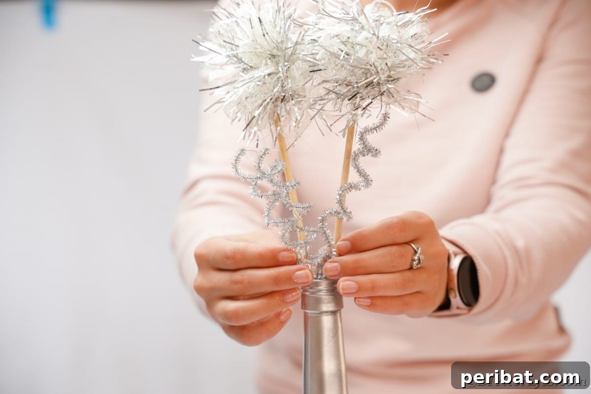 A person arranging tinsel-adorned dowels and pipe cleaner spirals into a silver wine bottle.