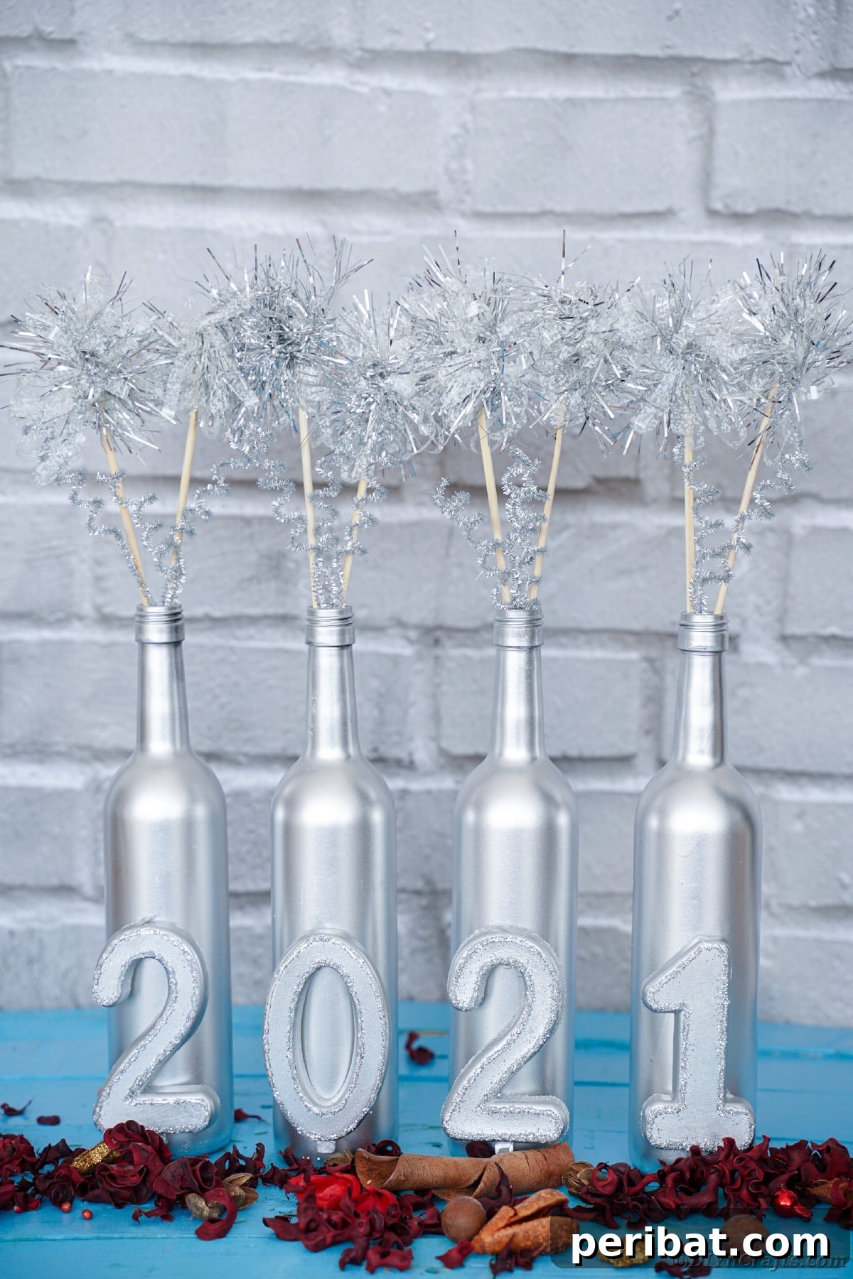 Silver-painted wine bottle decoration with numbers and tinsel accents, placed near a white brick wall.