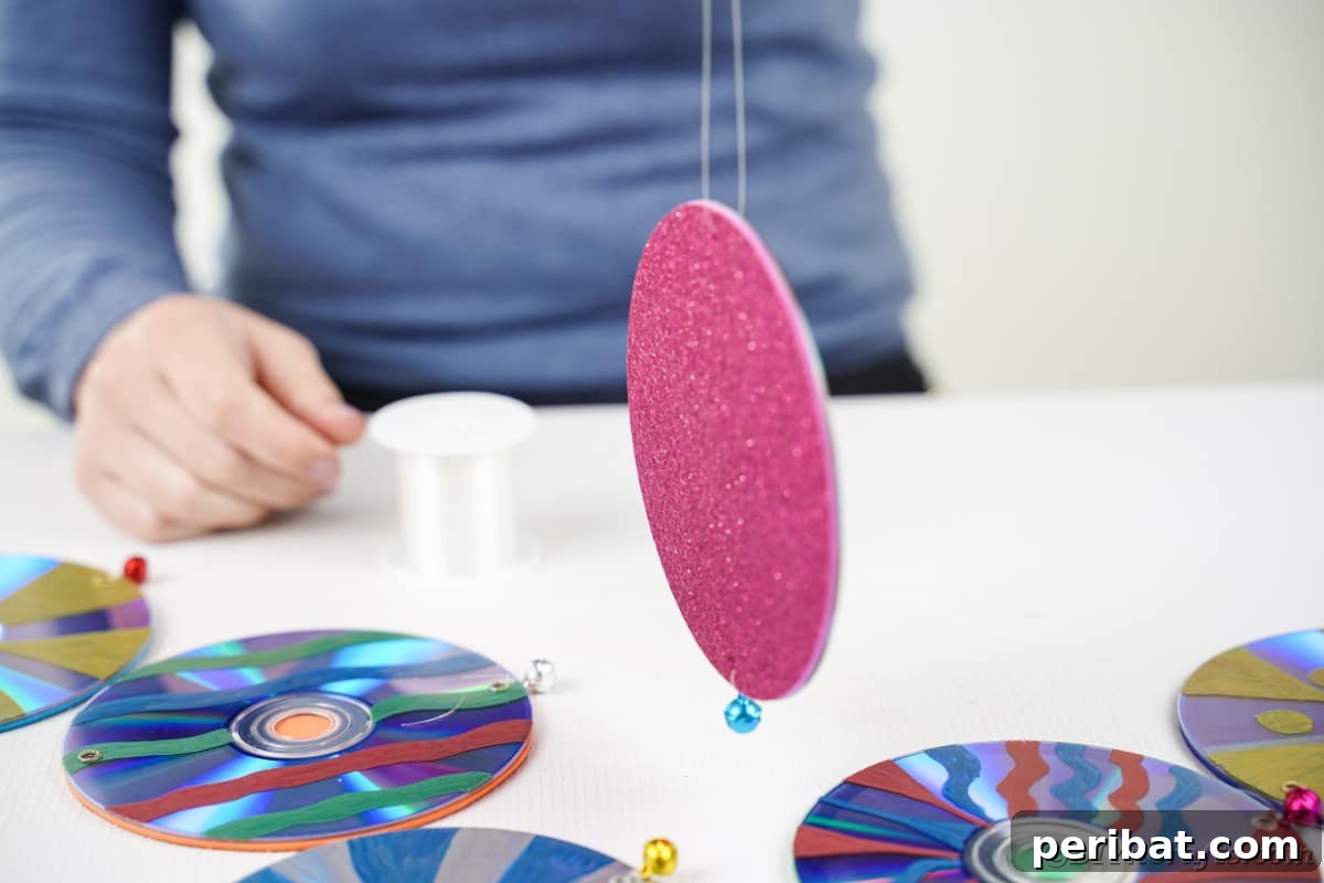 Sparkling Repurposed Garden Melody 17 A hand stringing a fishing line through a decorated CD, preparing to attach it to the main structure.
