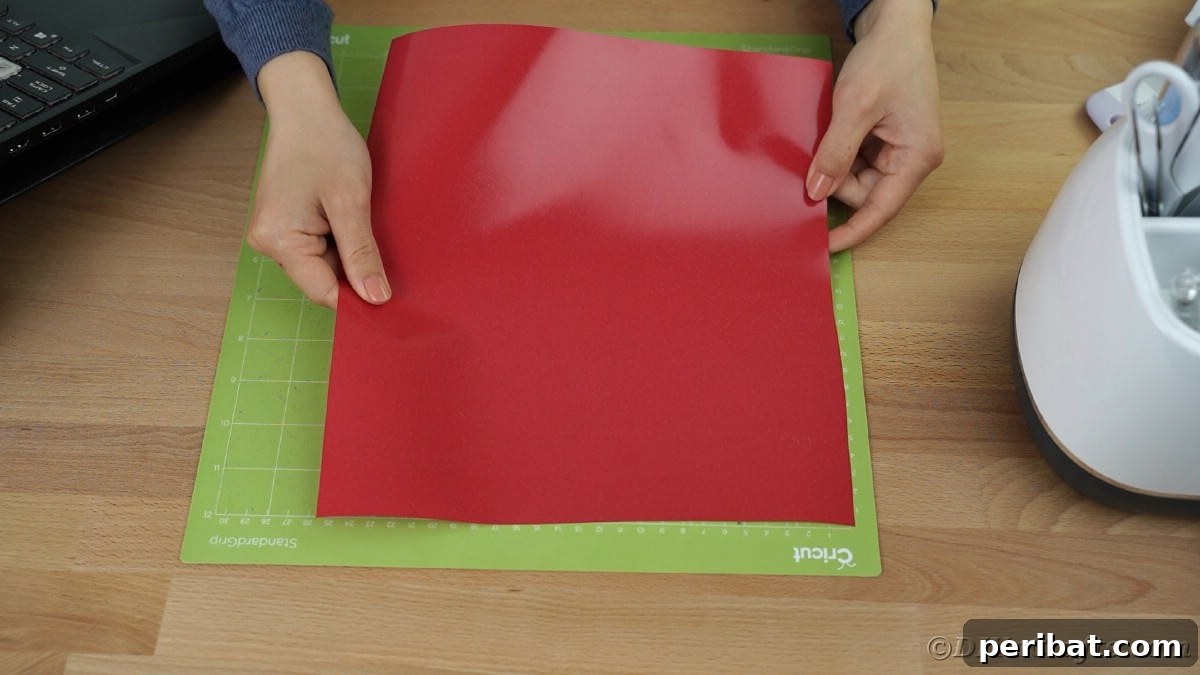 A hand carefully places a sheet of red heat transfer vinyl onto a green Cricut mat, ensuring the matte side is facing up and it's smooth.
