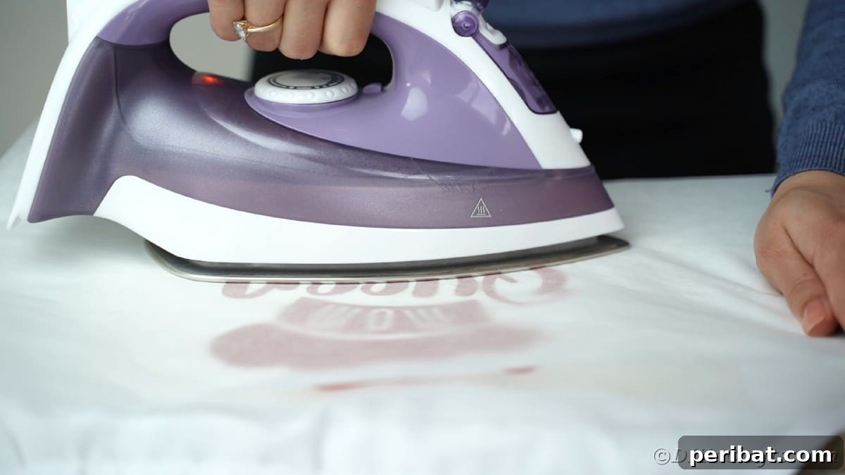 An iron is used to carefully apply heat over a piece of wax paper placed on a vinyl design, sealing it onto a white apron.
