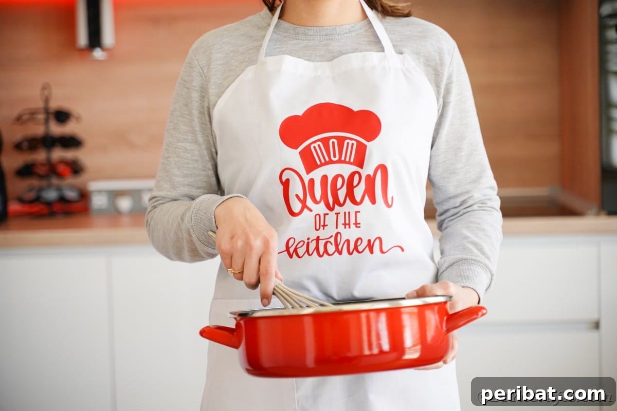 A woman proudly models a white and red Mother's Day apron with a custom Cricut vinyl design while holding a red skillet in a home kitchen setting.
