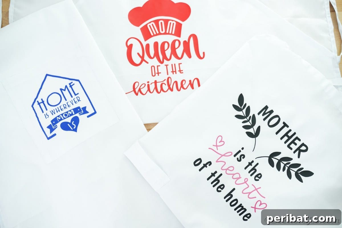 Three uniquely designed aprons, featuring red, blue, and black vinyl messages, are neatly stacked together on a soft pink surface, highlighting their individual styles.