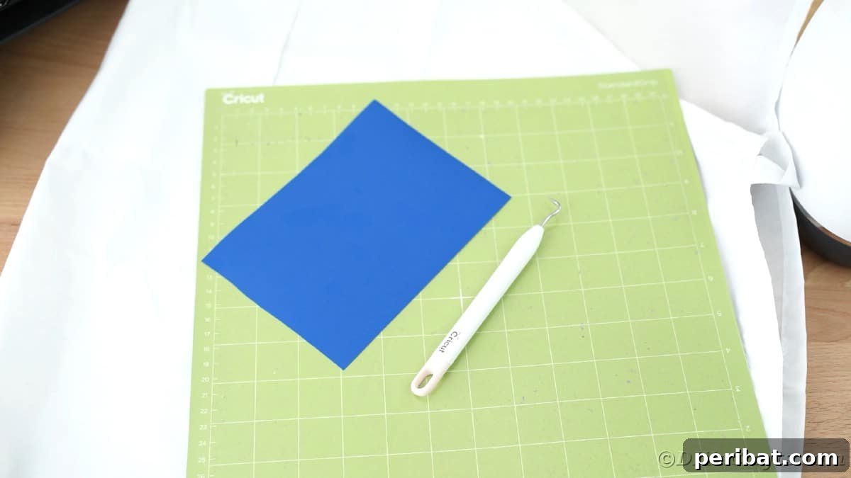 A blue heat transfer vinyl sheet is shown with a weeding tool, placed on a green Cricut cutting mat, ready for crafting the design.