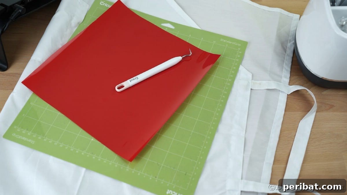 A white apron is displayed alongside a green Cricut cutting mat and a roll of red heat transfer vinyl, showcasing the essential tools for crafting.