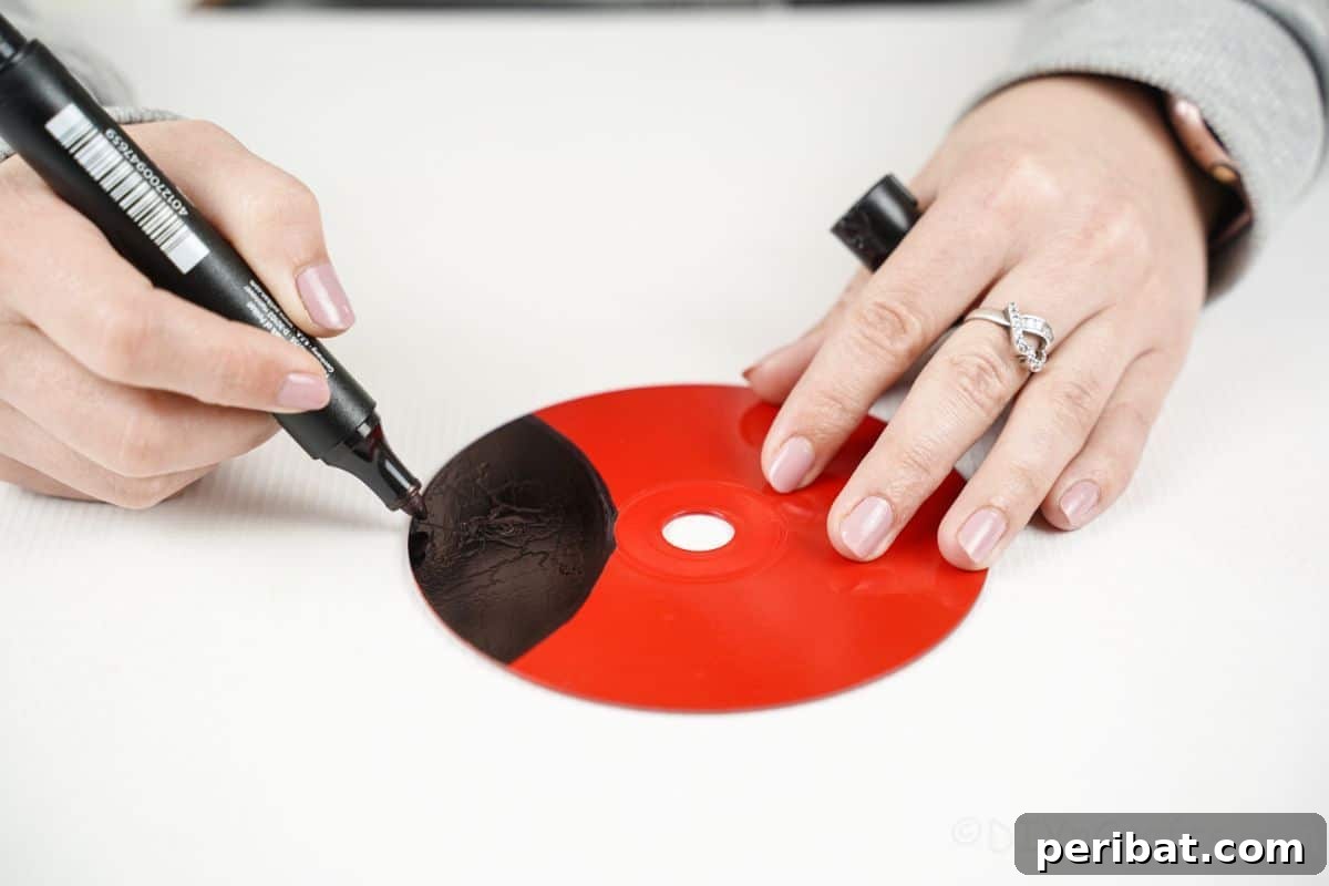 A hand holding a black marker, coloring in the designated head portion of the red-painted CD.