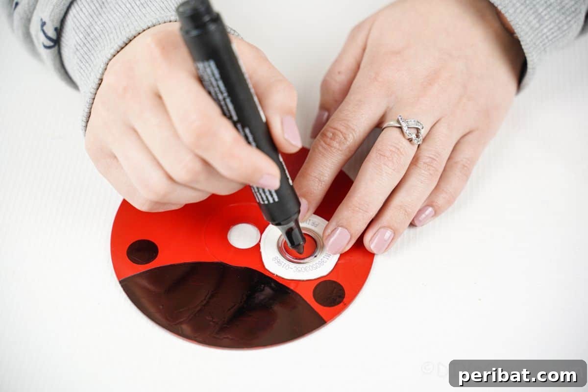 A hand holding a black permanent marker, tracing circles on the red-painted CD to create ladybug spots.