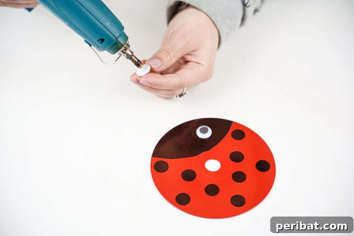 A hand applying craft glue to an upcycled CD ladybug, preparing to attach the googly eyes.