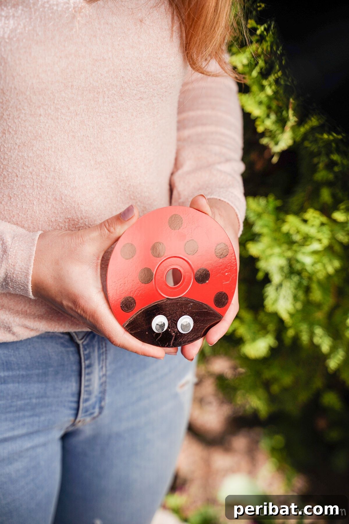 A woman in a pink top and jeans holding an upcycled CD ladybug, presenting the finished craft.