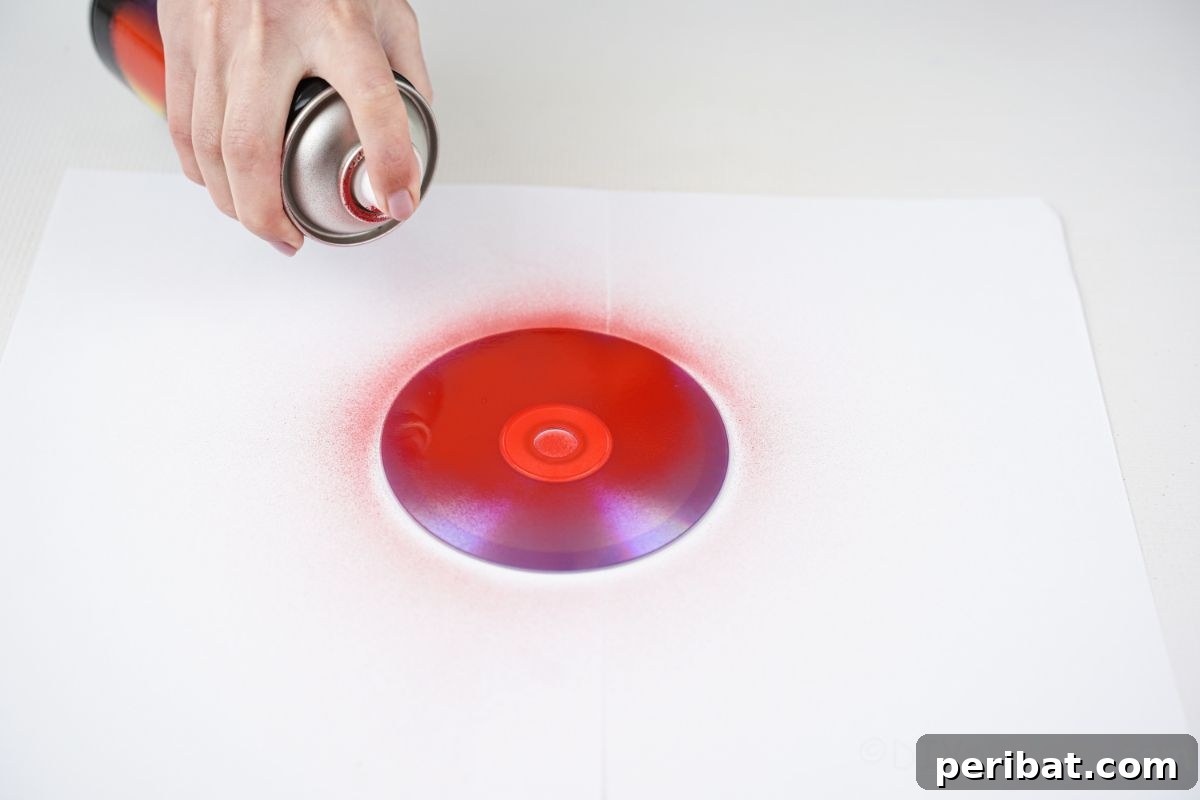 A hand spray painting an old CD red on a white table, demonstrating the first step of the craft.