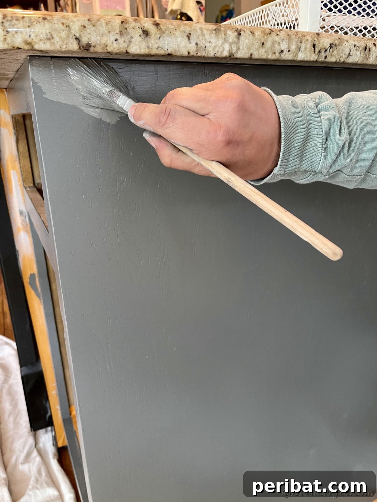 A hand painting a cabinet with a brush, applying the second coat of gray paint