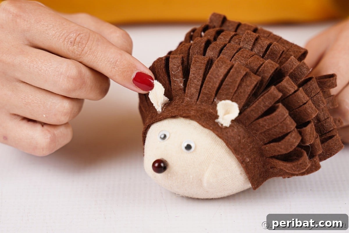 Effortless No-Stitch Fabric Hedgehog Creation 18 Hands carefully glue small, oval-shaped fabric pieces onto the top of the hedgehog's head, forming its adorable ears.