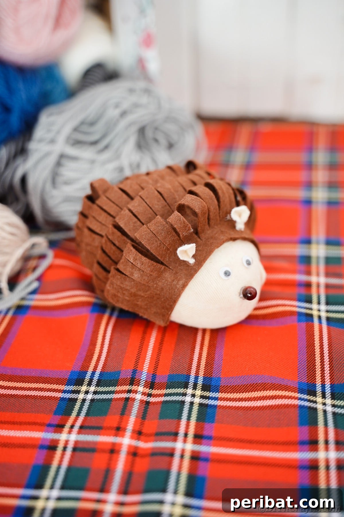 Effortless No-Stitch Fabric Hedgehog Creation 3 A charming stuffed toy hedgehog rests on a cozy red plaid blanket, surrounded by rolls of soft yarn, evoking a warm, autumnal feeling.