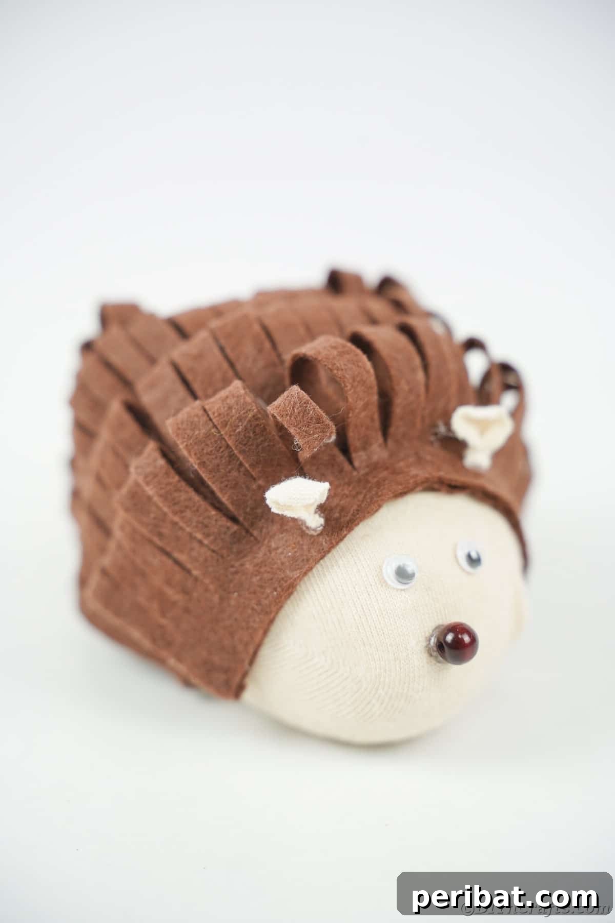 Effortless No-Stitch Fabric Hedgehog Creation 20 A pristine white background serves as a minimalist setting for a charming stuffed fabric hedgehog, highlighting its texture and form.
