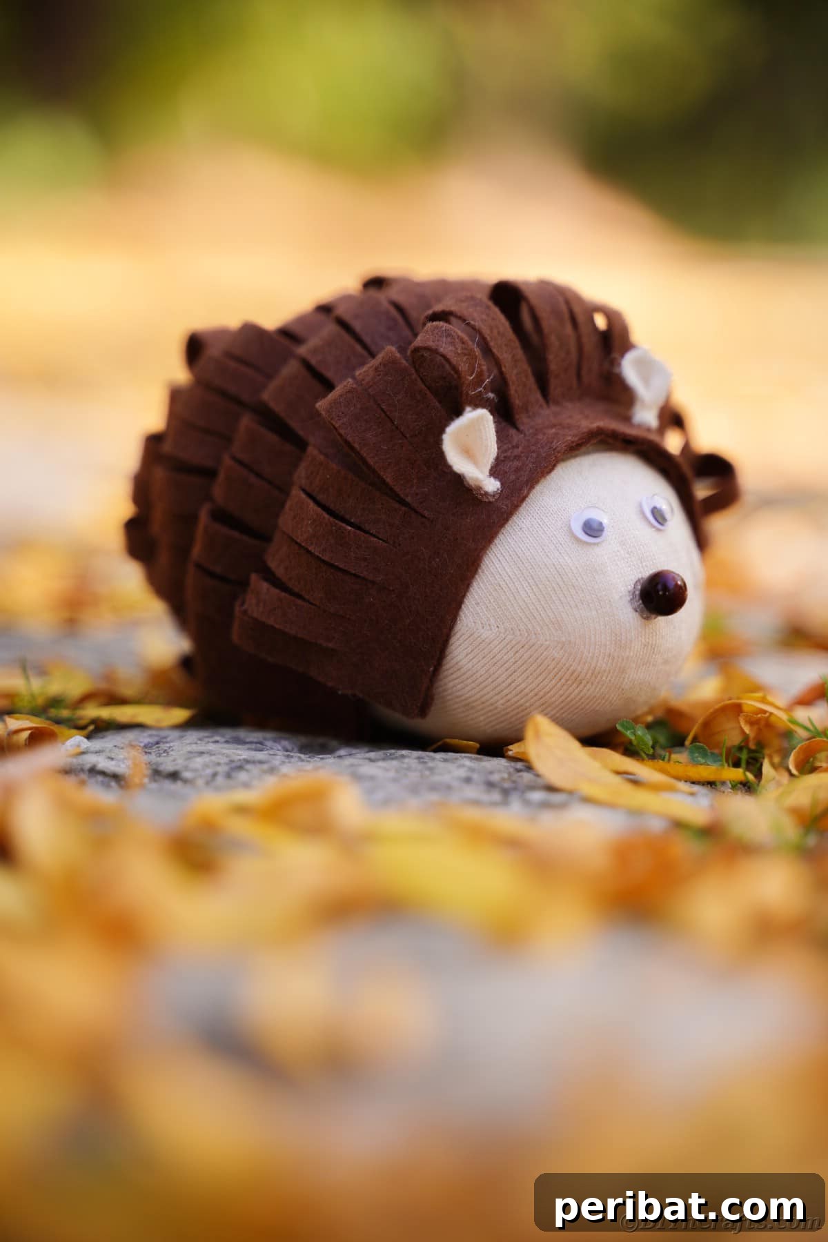 Effortless No-Stitch Fabric Hedgehog Creation 9 A sweet stuffed toy hedgehog sits on the ground outdoors, surrounded by a scattering of vibrant autumn leaves, embodying a whimsical woodland scene.