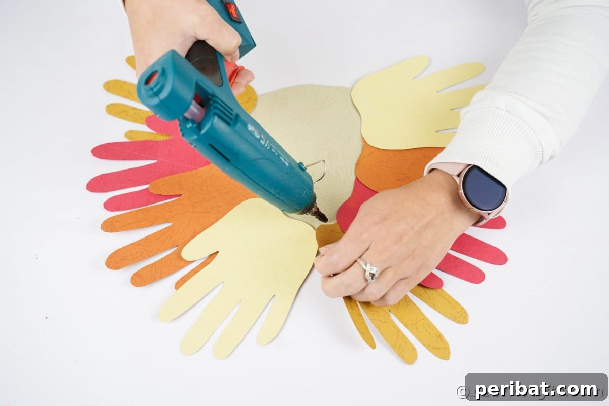 A hand holding a blue hot glue gun, carefully applying glue to a paper handprint before attaching it to the hedgehog body