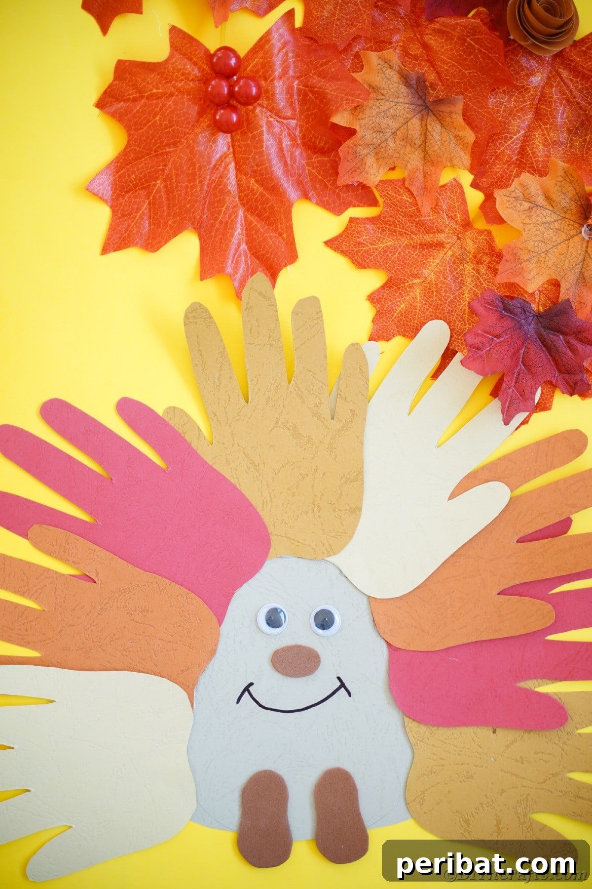 Finished handprint hedgehog craft resting on a bright yellow background adorned with faux autumn leaves