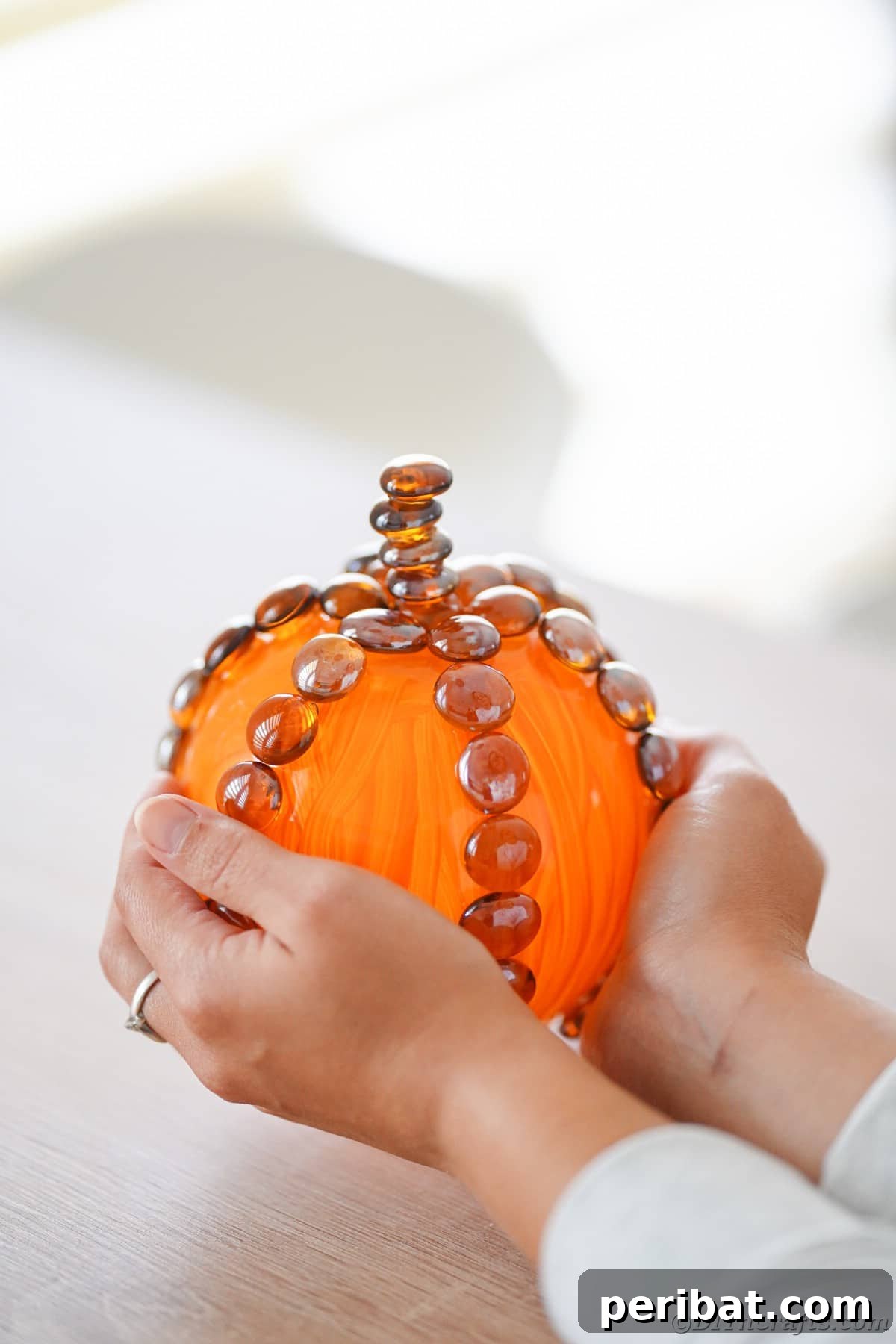 Hands holding a beautifully painted glass pumpkin, showcasing its autumn charm on a rustic table.